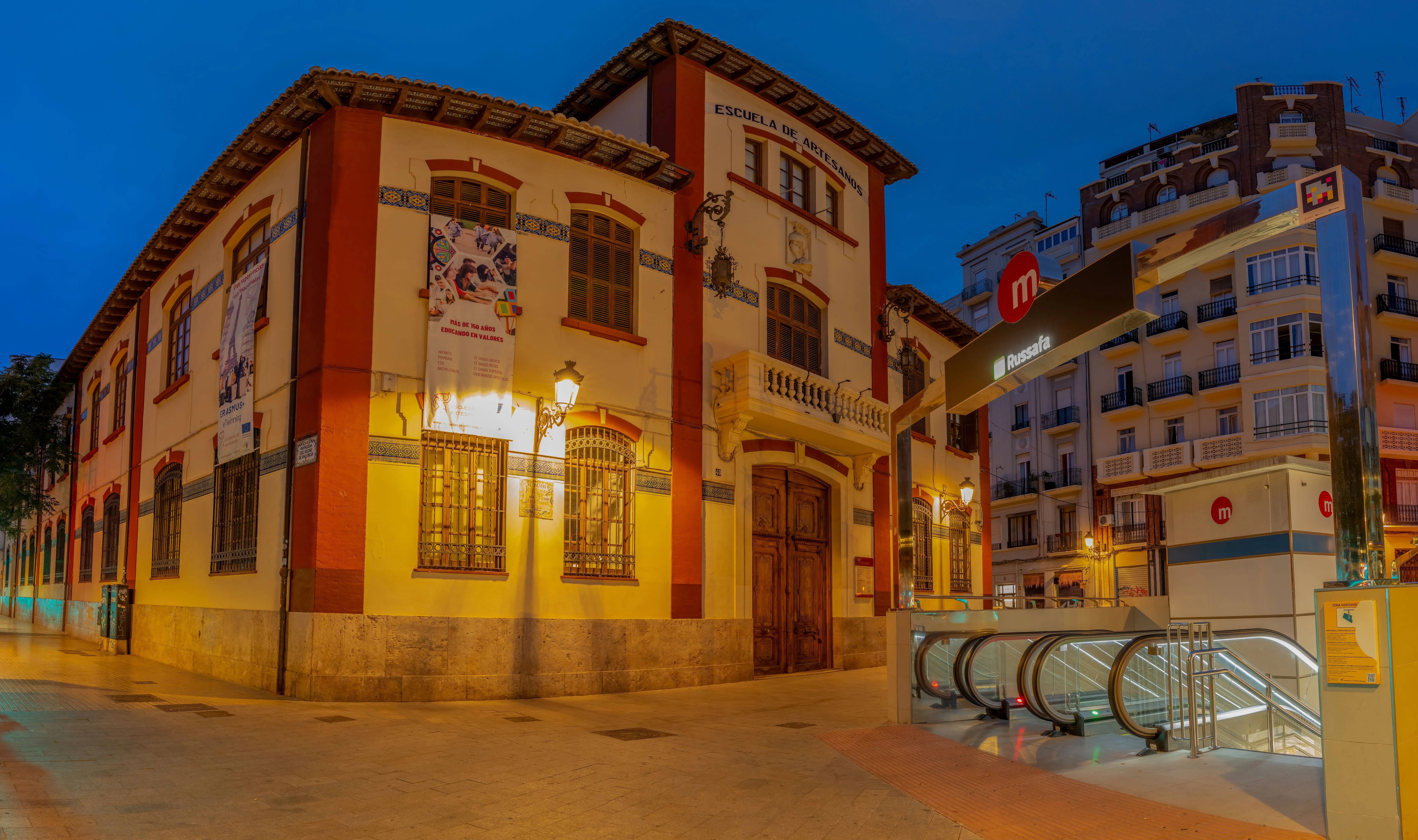 The old Escuela de Artesanos building, ochre with red-brick trim, beside the entrance to Russafa metro station at dusk, with blocks of flats behind it.